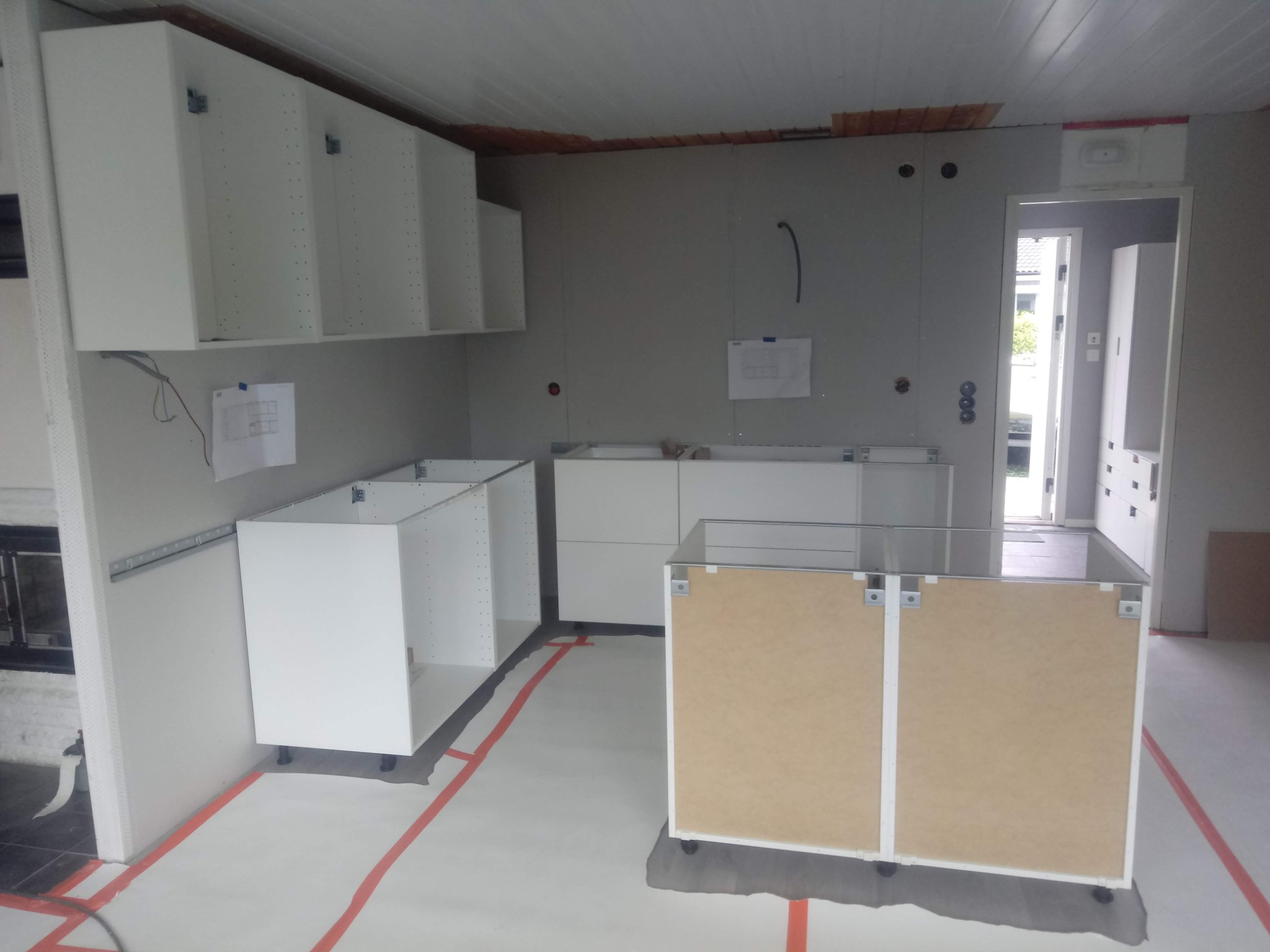 Montaż białych szafek kuchennych: widoczne górne i dolne moduły, zabezpieczona podłoga, surowe ściany przygotowane do wykończenia, widok na inne pomieszczenie w tle.