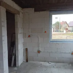 Surowe wnętrze w trakcie budowy z widoczną instalacją elektryczną: białe kable prowadzone po ścianach z bloków, pomarańczowe puszki elektryczne, okno z widokiem na domy.