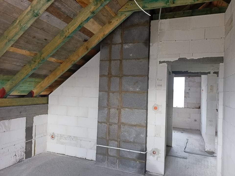 Surowy stan poddasza z widoczną instalacją elektryczną: przewody prowadzone po ścianie z bloczków, puszka instalacyjna z pomarańczową pokrywką, widoczna konstrukcja dachu.