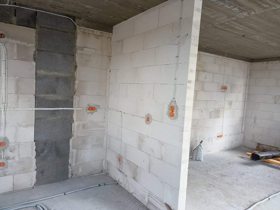 Wnętrze w trakcie instalacji elektrycznej: widoczne ściany z betonu komórkowego, puszki elektryczne w kolorze pomarańczowym i przewody rozprowadzone po ścianach, surowy betonowy strop.