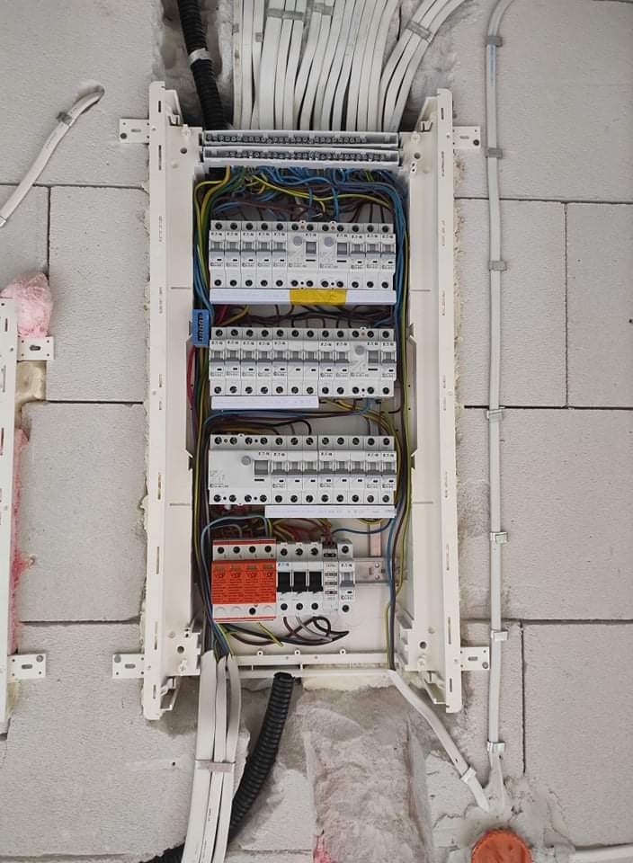 Rozdzielnica elektryczna w trakcie montażu w surowej ścianie z betonu komórkowego, widoczne kable, bezpieczniki i ogranicznik przepięć.