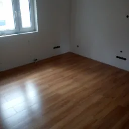 Pokój po położeniu paneli podłogowych w odcieniach drewna, widoczne gniazdka elektryczne na białych ścianach i okno z białymi ramami.