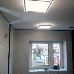 Wykończona łazienka z marmurowymi płytkami, okno z widokiem na zewnątrz i dwie nowoczesne, kwadratowe lampy sufitowe z białym światłem.