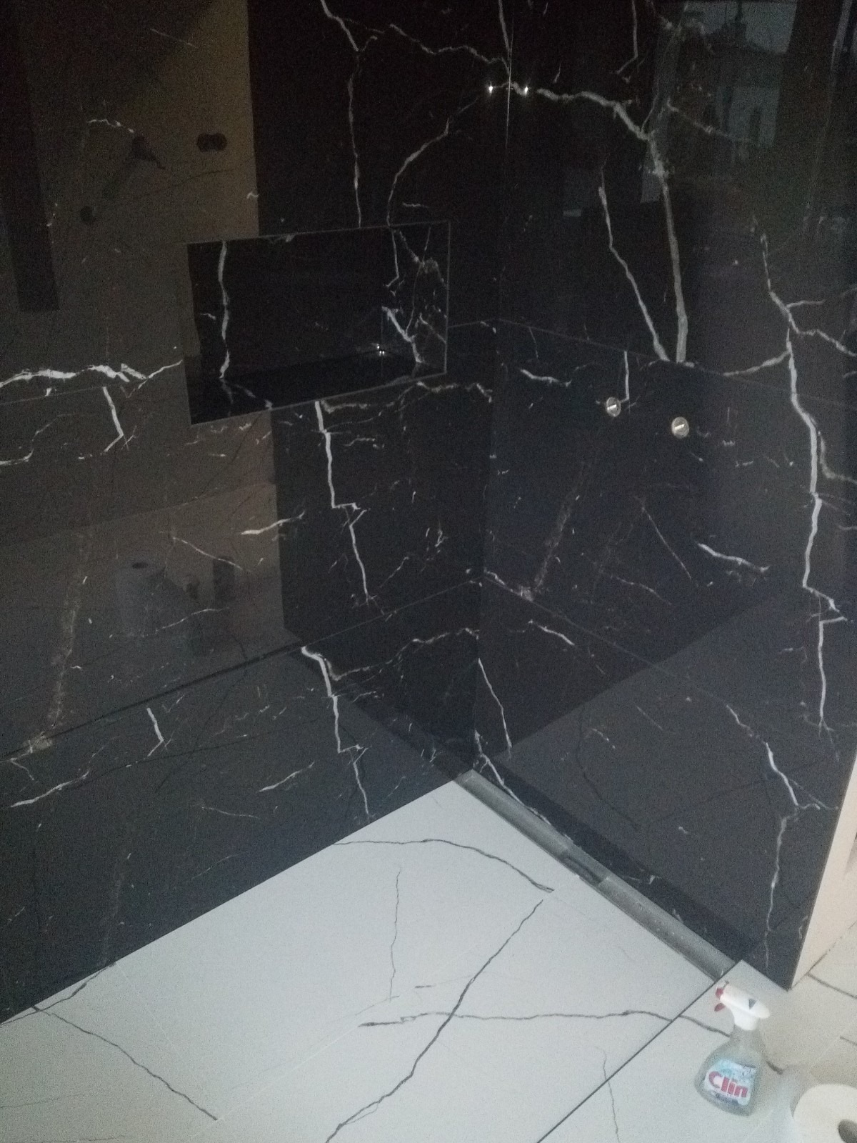 Wykończona kabina prysznicowa z czarnymi płytkami imitującymi marmur z białymi żyłami, z wbudowaną półką i dwoma punktowymi lampami, na podłodze białe płytki z szarymi żyłami, widoczna butelka...