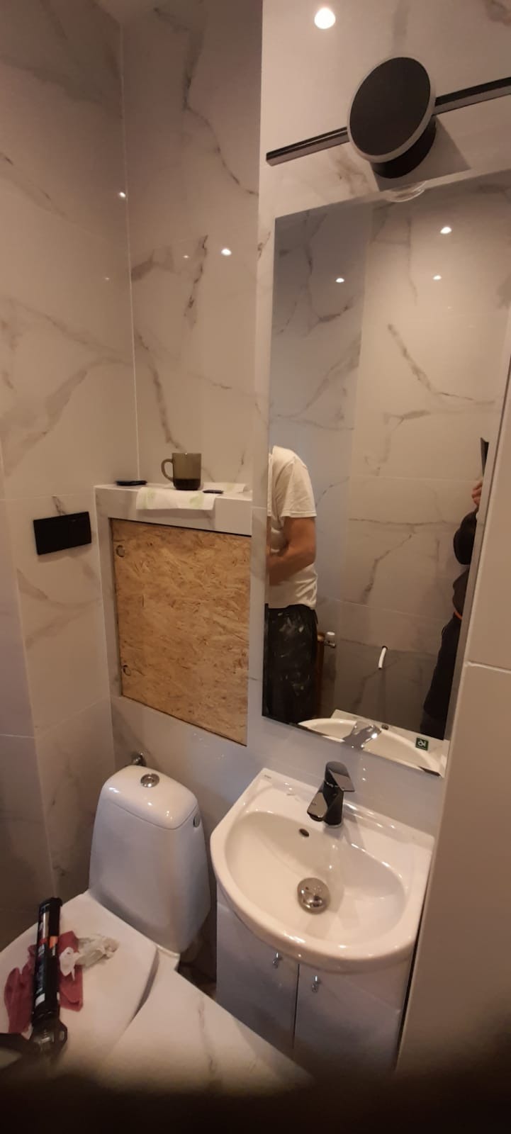 Wnętrze łazienki w trakcie prac: białe płytki imitujące marmur, toaleta, umywalka z czarną baterią, lustro, zakryty otwór rewizyjny płytą OSB, widoczny pracownik w odbiciu lustra.