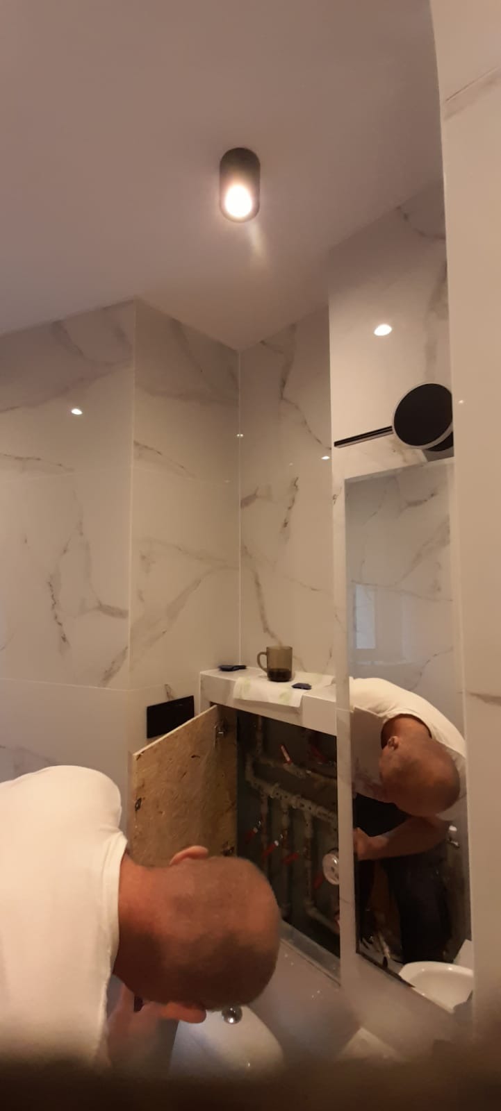 Hydraulik w trakcie montażu instalacji pod umywalką w łazience z marmurowymi płytkami, widoczne otwarte drzwiczki szafki i kubek na blacie.