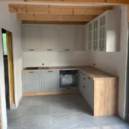 Wnętrze kuchni w trakcie wykańczania: szare szafki z drewnianym blatem, drewniany sufit i fragment ściany z boazerią, szare płytki podłogowe, widok na zewnątrz przez okno.