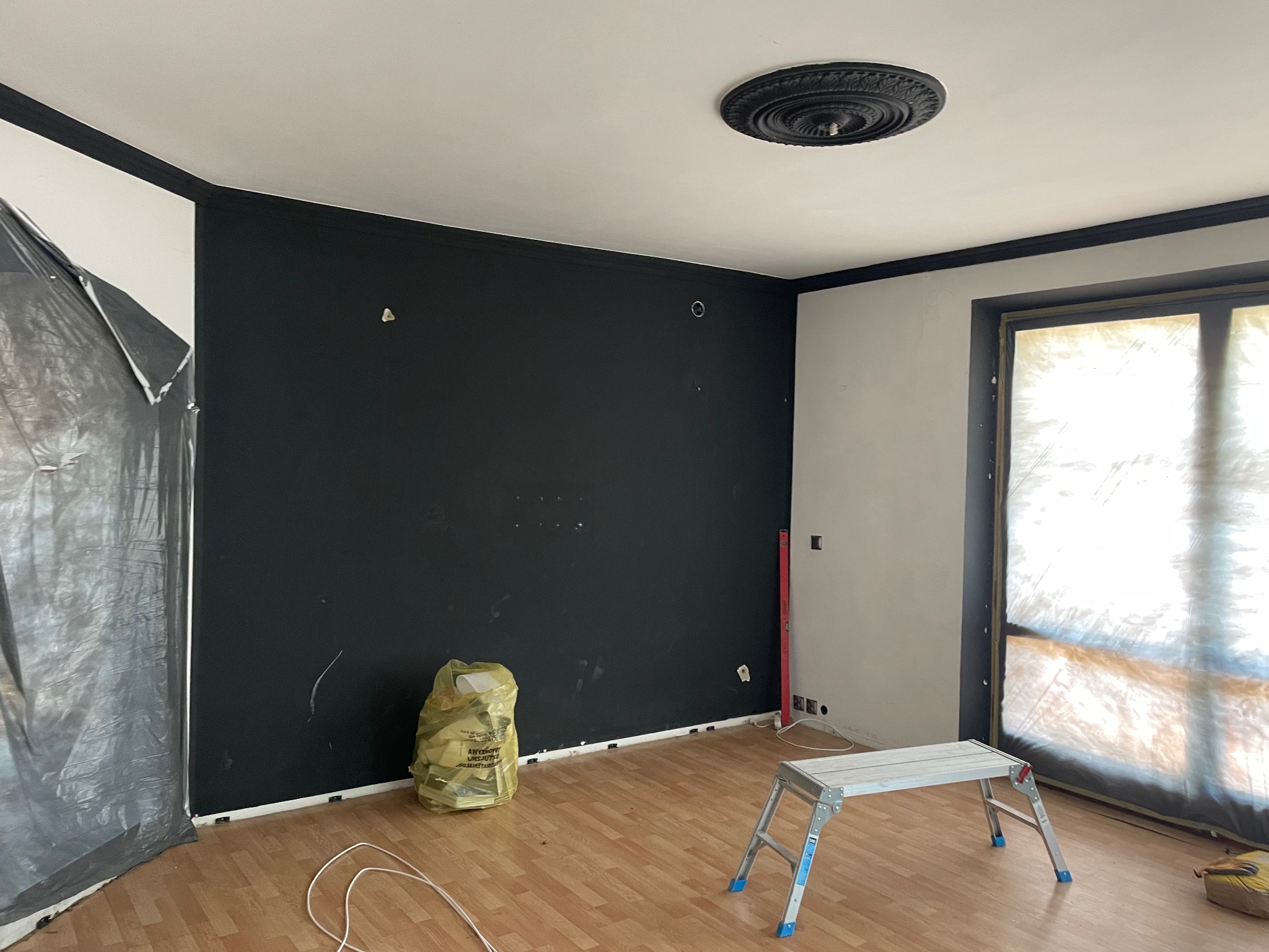 Pokój w trakcie odświeżania: ściana pomalowana na czarno, zabezpieczone okno folią malarską, drabina aluminiowa, worek z odpadami i listwa przypodłogowa w kolorze białym.