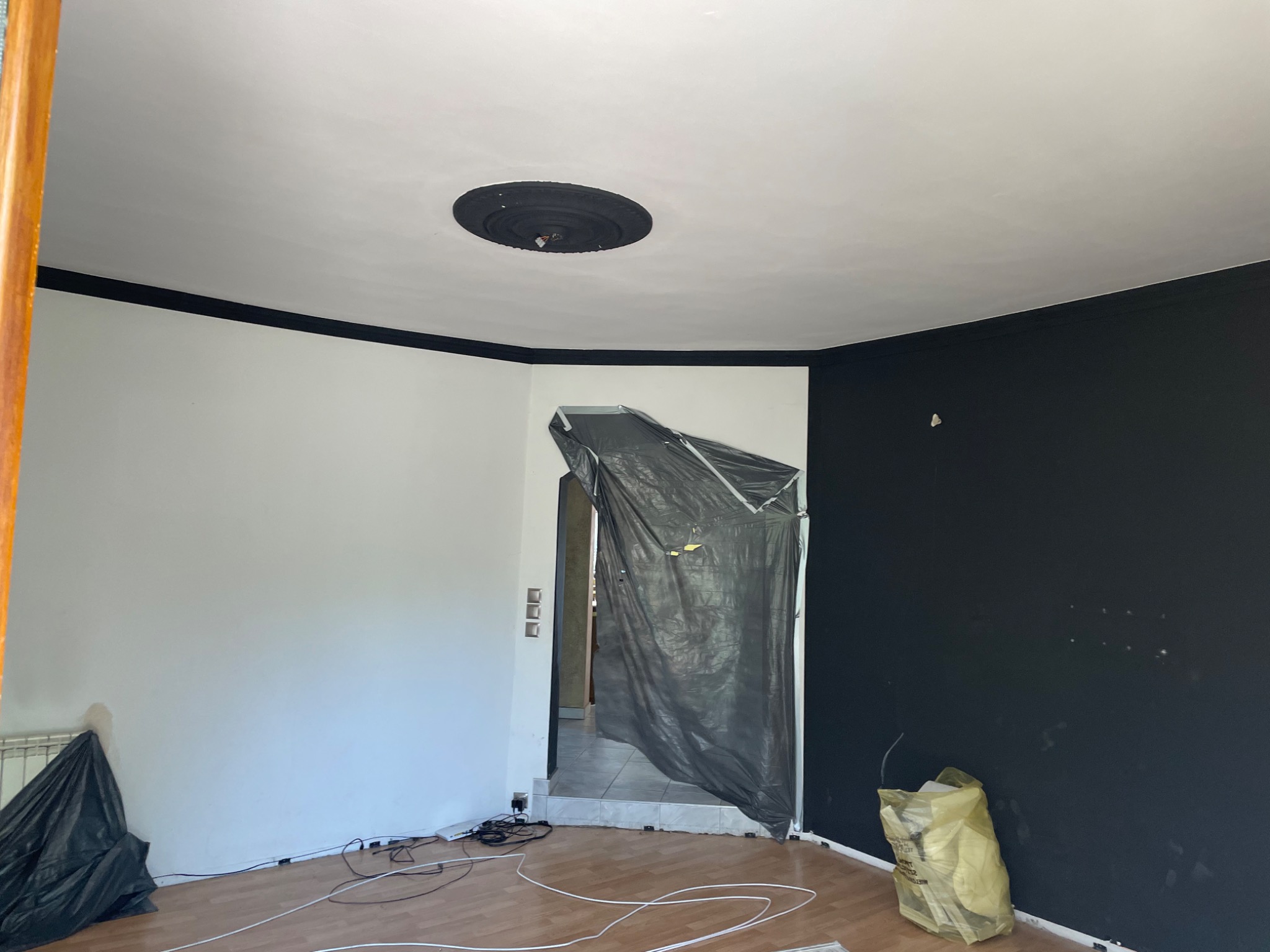 Pokój w trakcie malowania: jedna ściana biała, druga czarna, z czarnym paskiem pod sufitem, zabezpieczone folią drzwi, widoczne kable i worki na podłodze.