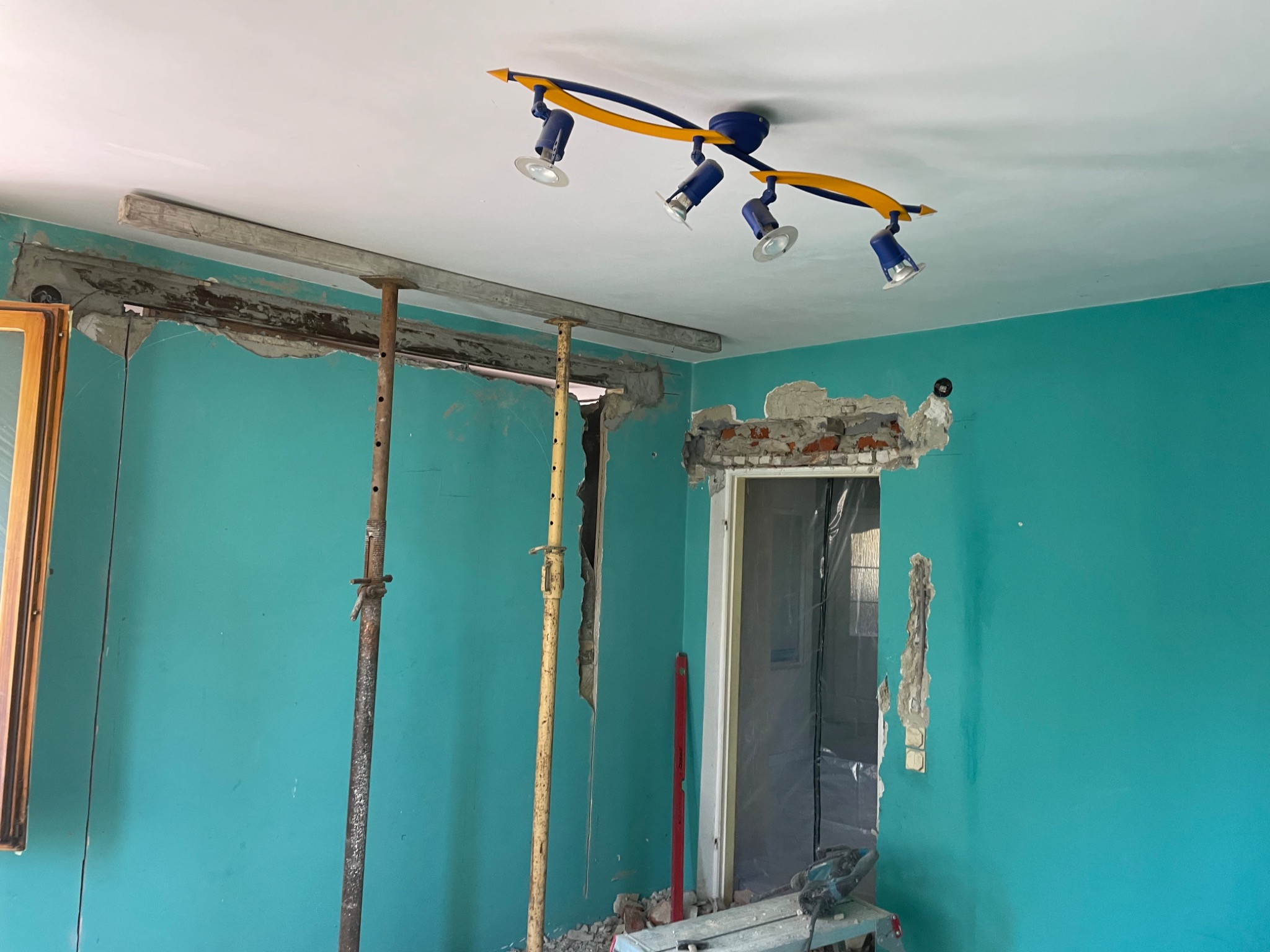Wnętrze w trakcie prac remontowych, widoczne podparte belki stropowe, otwór w ścianie, niebieska ściana, lampa sufitowa z reflektorami i narzędzia na podłodze.