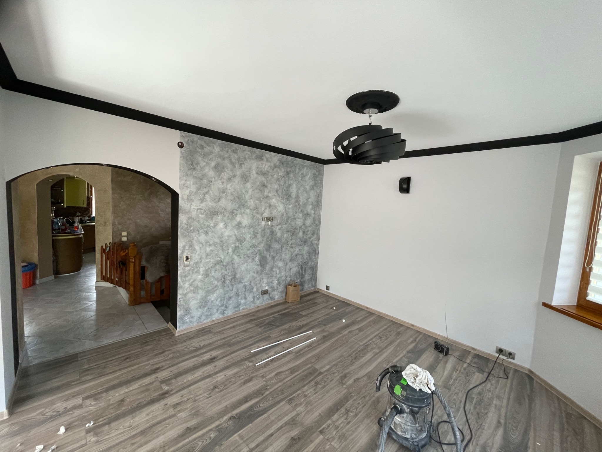 Pokój po remoncie z panelami podłogowymi, ścianą z efektem dekoracyjnym i czarnym wykończeniem sufitu, widoczny odkurzacz przemysłowy i elementy wykończeniowe na podłodze.