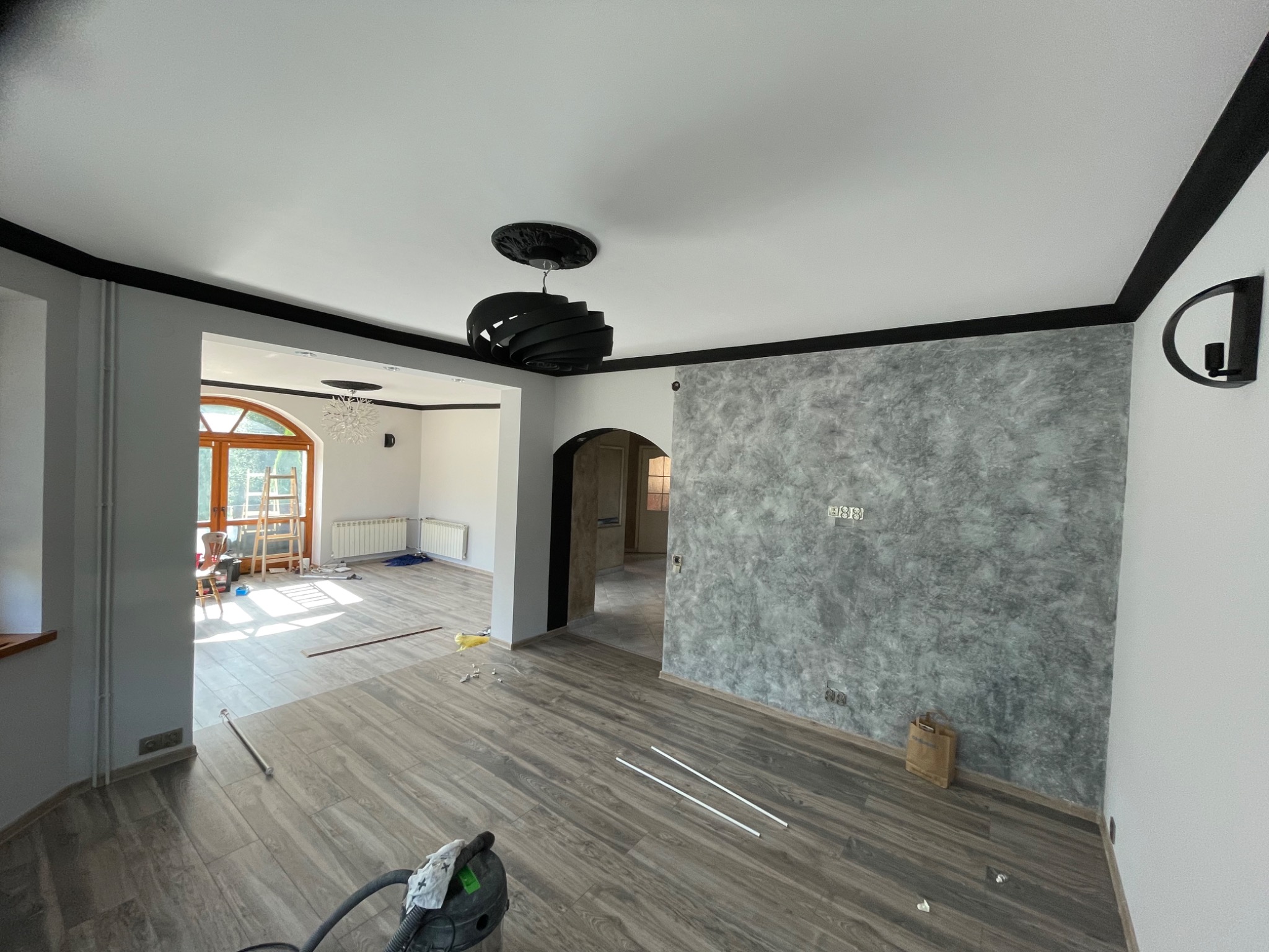 Wnętrze w trakcie prac wykończeniowych: położona podłoga z paneli imitujących drewno, ściana z efektem dekoracyjnym w odcieniach szarości, widoczne listwy przypodłogowe i odkurzacz, sufit...