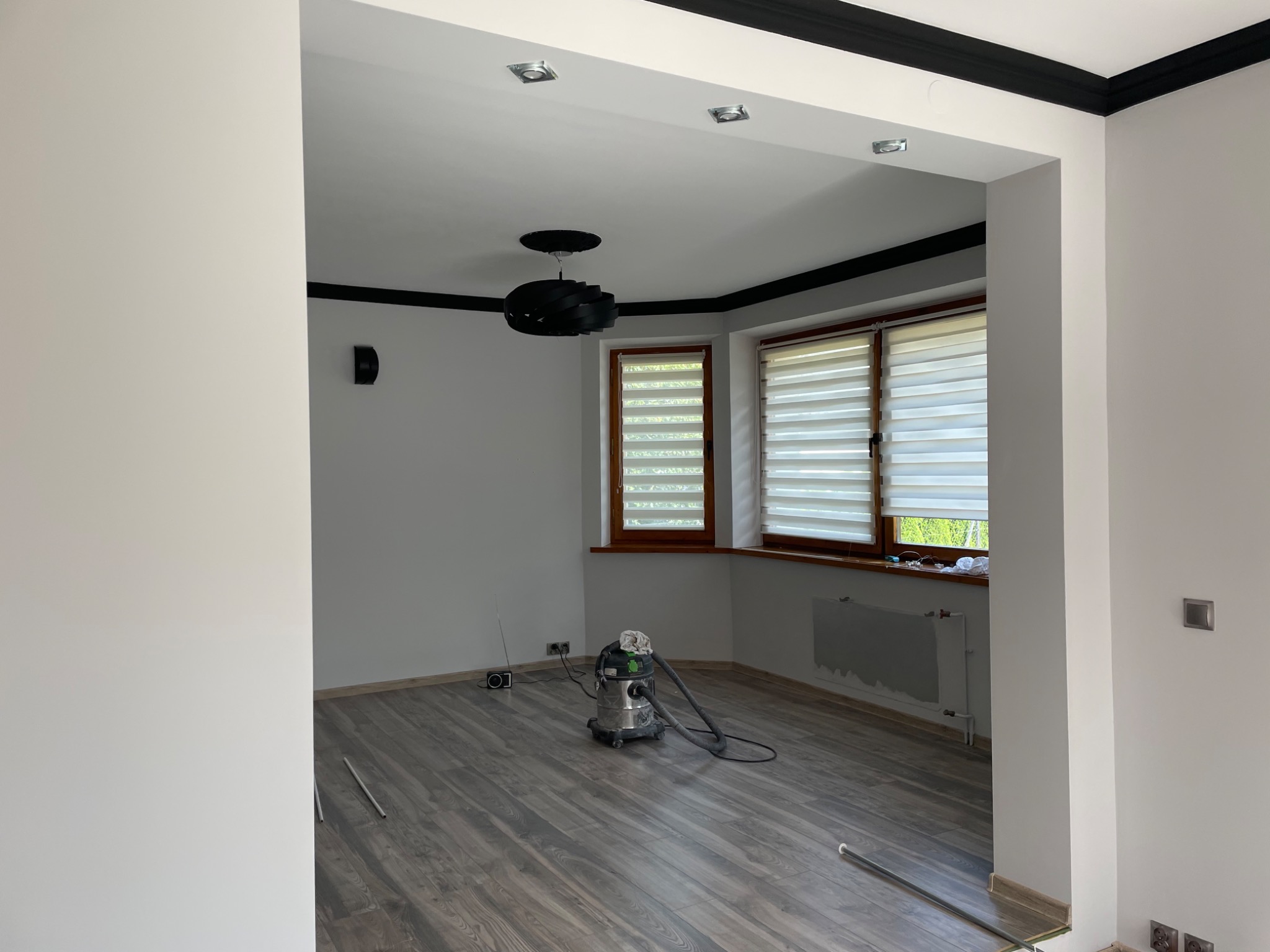 Pokój po remoncie z szarymi panelami podłogowymi, świeżo pomalowanymi ścianami na biało z czarnymi listwami przysufitowymi, nowoczesną lampą i odkurzaczem przemysłowym na środku