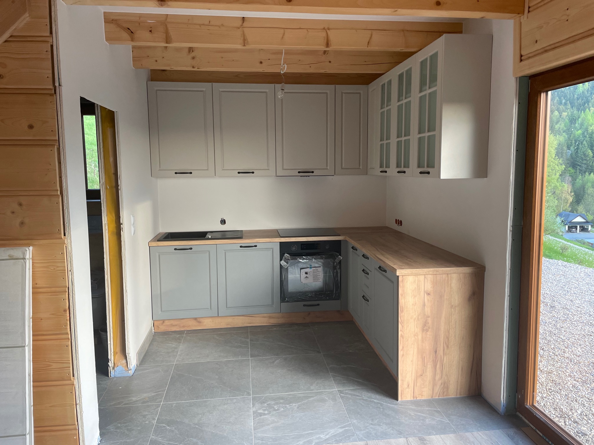 Wnętrze kuchni w trakcie wykańczania: szare szafki z drewnianym blatem, drewniany sufit i fragment ściany z boazerią, szare płytki podłogowe, widok na zewnątrz przez okno.