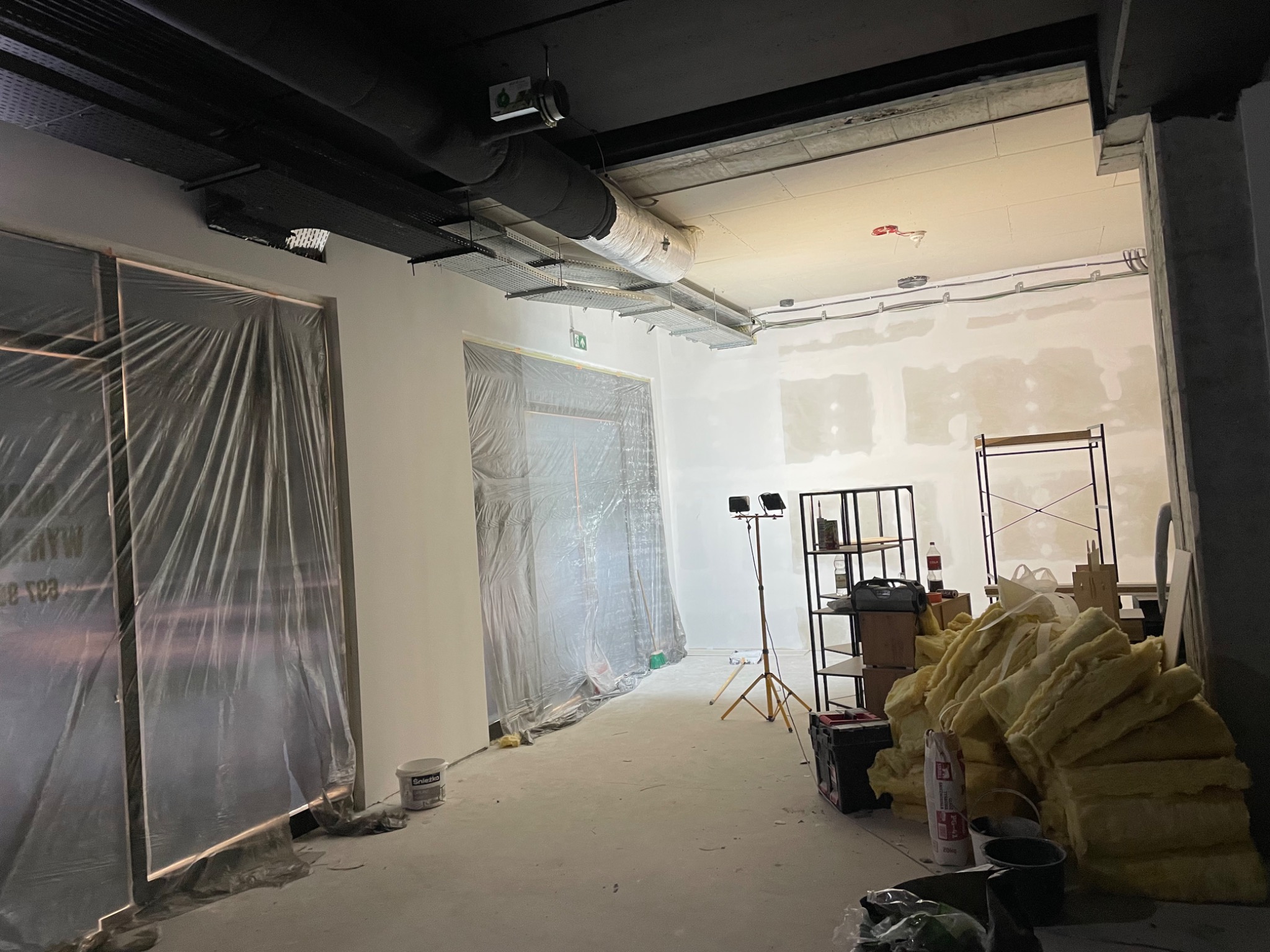 Wnętrze w trakcie prac wykończeniowych: ściany przygotowane pod gładzie, zabezpieczone folią drzwi, widoczne materiały budowlane i oświetlenie robocze.