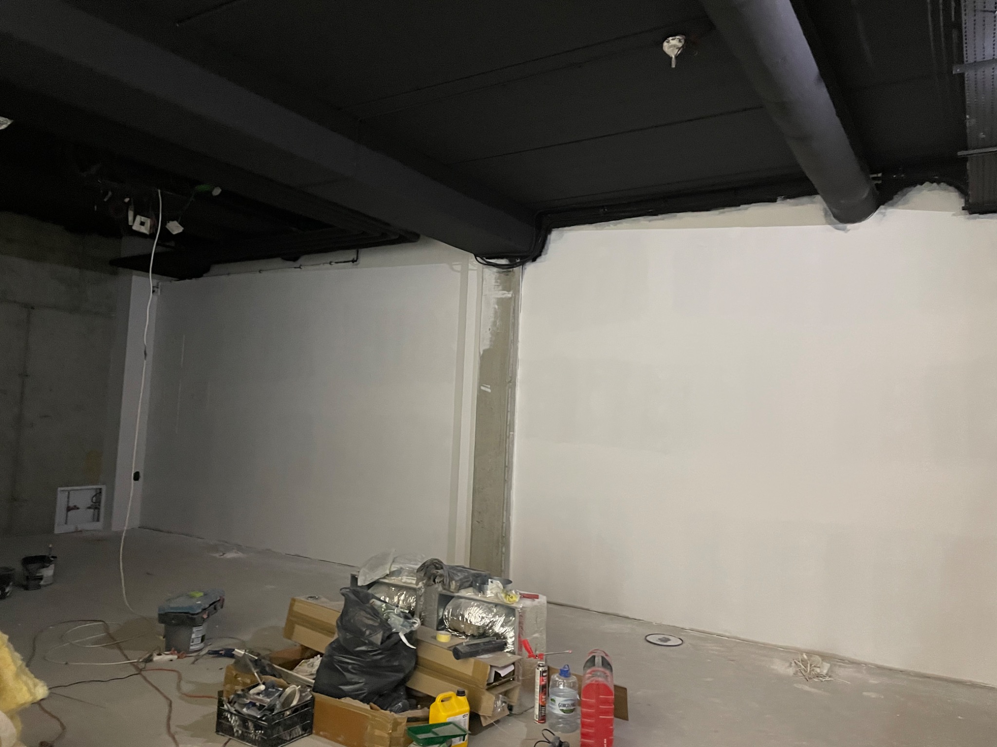 Wnętrze w trakcie prac wykończeniowych: białe ściany, czarny sufit z instalacjami, widoczne narzędzia i materiały budowlane. Ściany świeżo pomalowane, na podłodze bałagan związany z prowadzonymi...