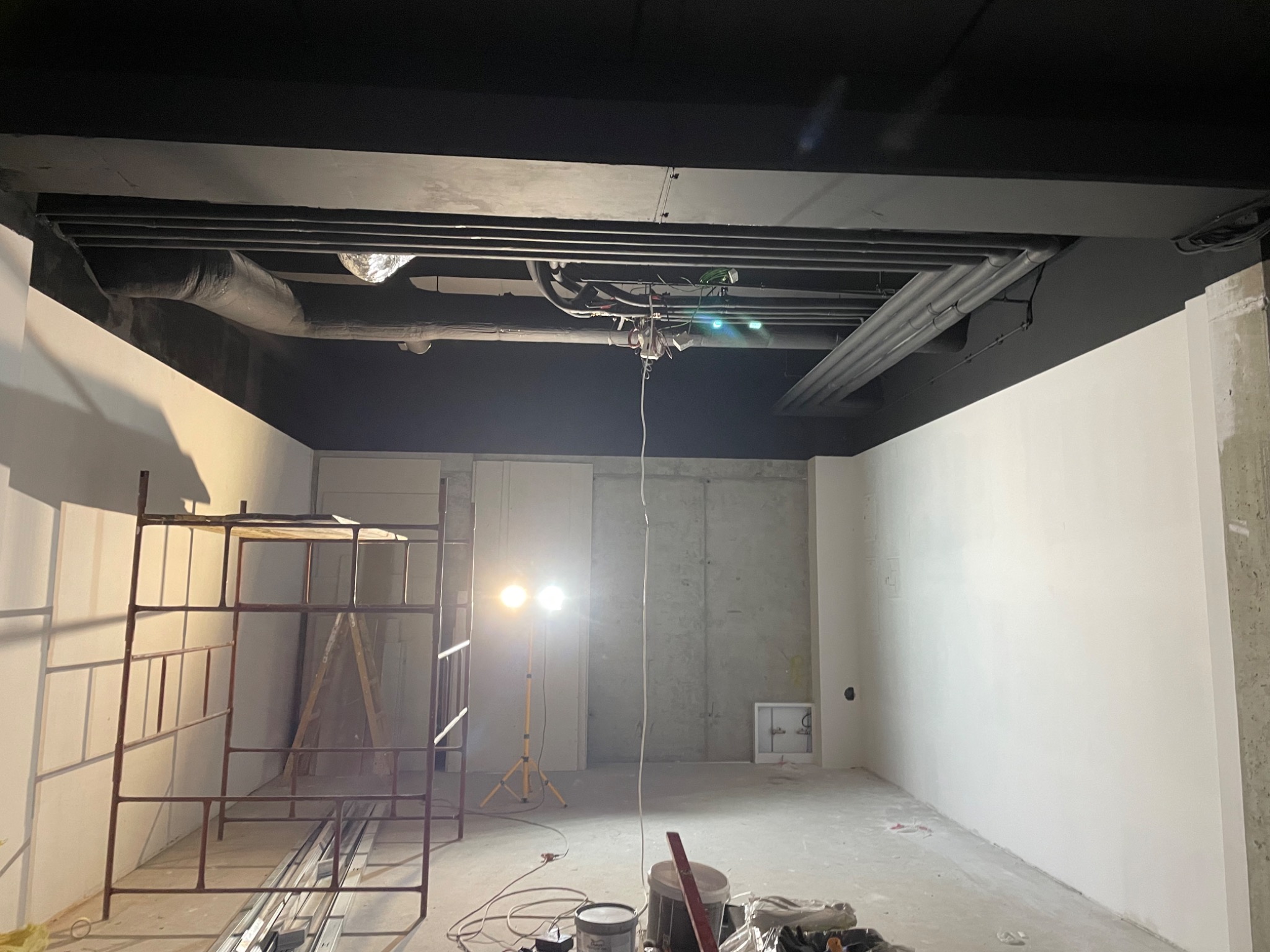 Wnętrze w trakcie prac, widoczne rusztowanie, lampa budowlana, puszki z farbą i poziomica, sufit pomalowany na czarno, ściany częściowo pomalowane na biało, instalacje pod sufitem.