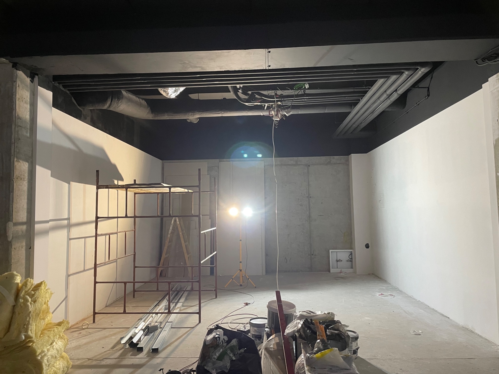 Wnętrze w trakcie prac wykończeniowych: ściany częściowo pomalowane na biało, widoczne rusztowanie, oświetlenie robocze, materiały izolacyjne i narzędzia na podłodze, instalacje pod sufitem.