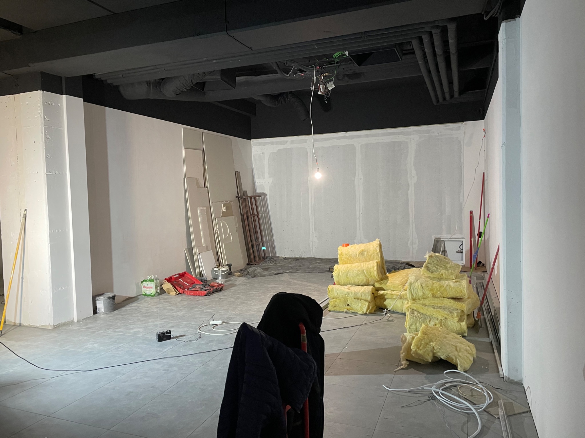 Wnętrze w trakcie prac, widoczne płyty gipsowo-kartonowe na ścianach, izolacja z wełny mineralnej, narzędzia i materiały budowlane rozłożone na podłodze z szarych płytek, surowy stan wykończenia.