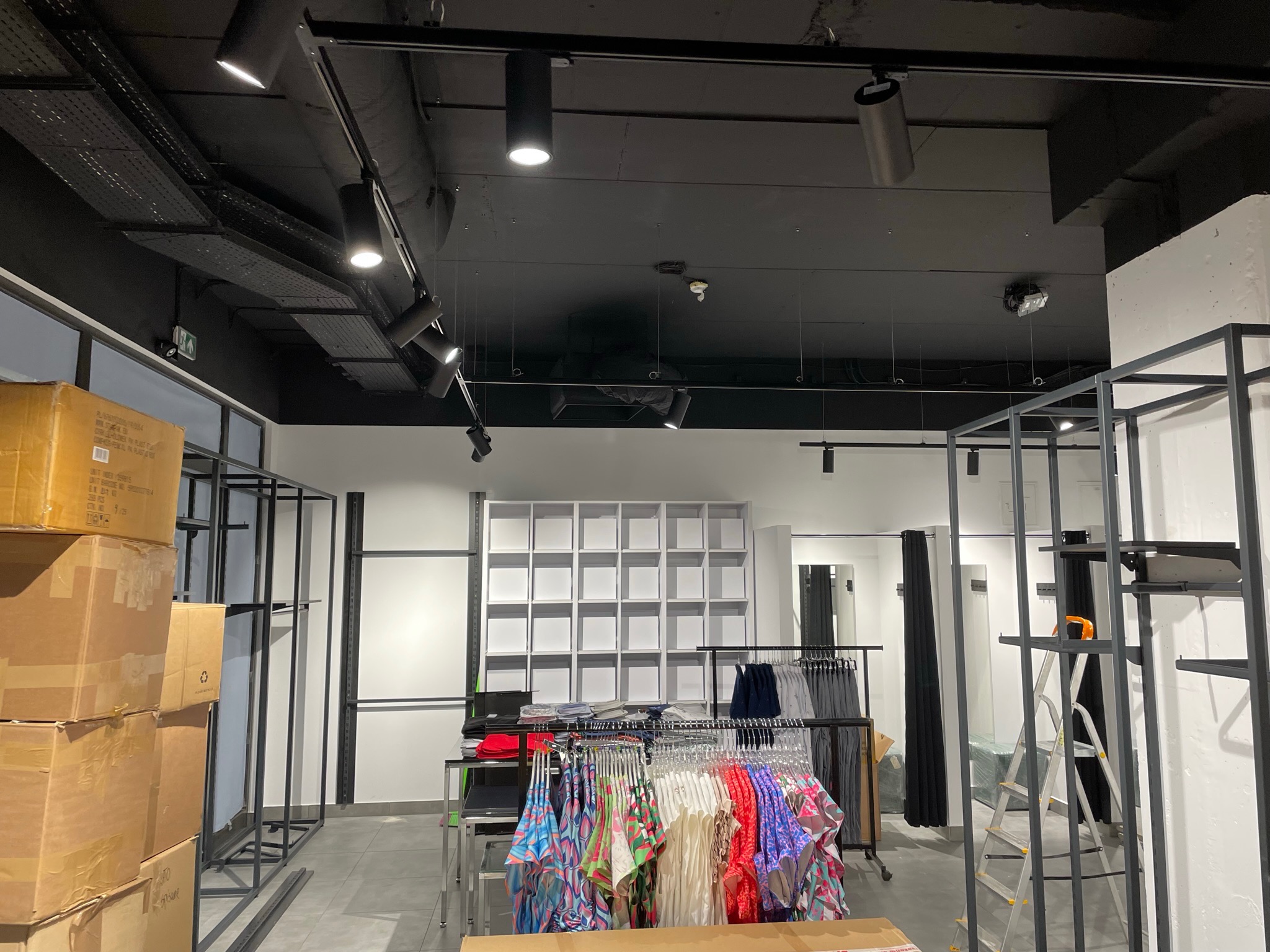 Wnętrze sklepu odzieżowego w trakcie aranżacji: widoczne metalowe regały, kartony z towarem, przymierzalnie z zasłonami i oświetlenie sufitowe.