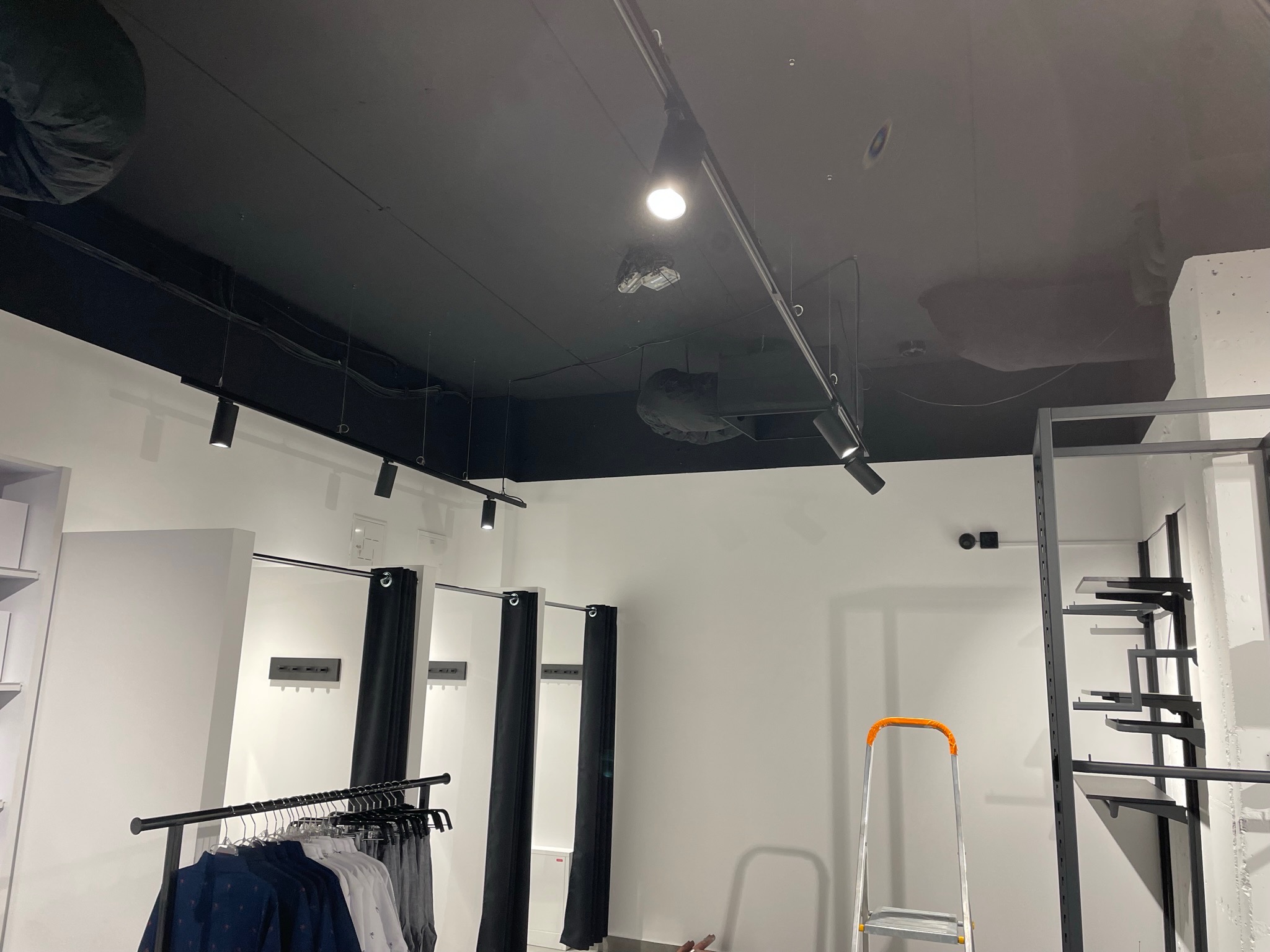 Wnętrze sklepu odzieżowego w trakcie budowy lub po remoncie, widoczny czarny sufit z oświetleniem szynowym, białe ściany, przymierzalnie z czarnymi zasłonami, drabina i metalowe regały bez towaru.