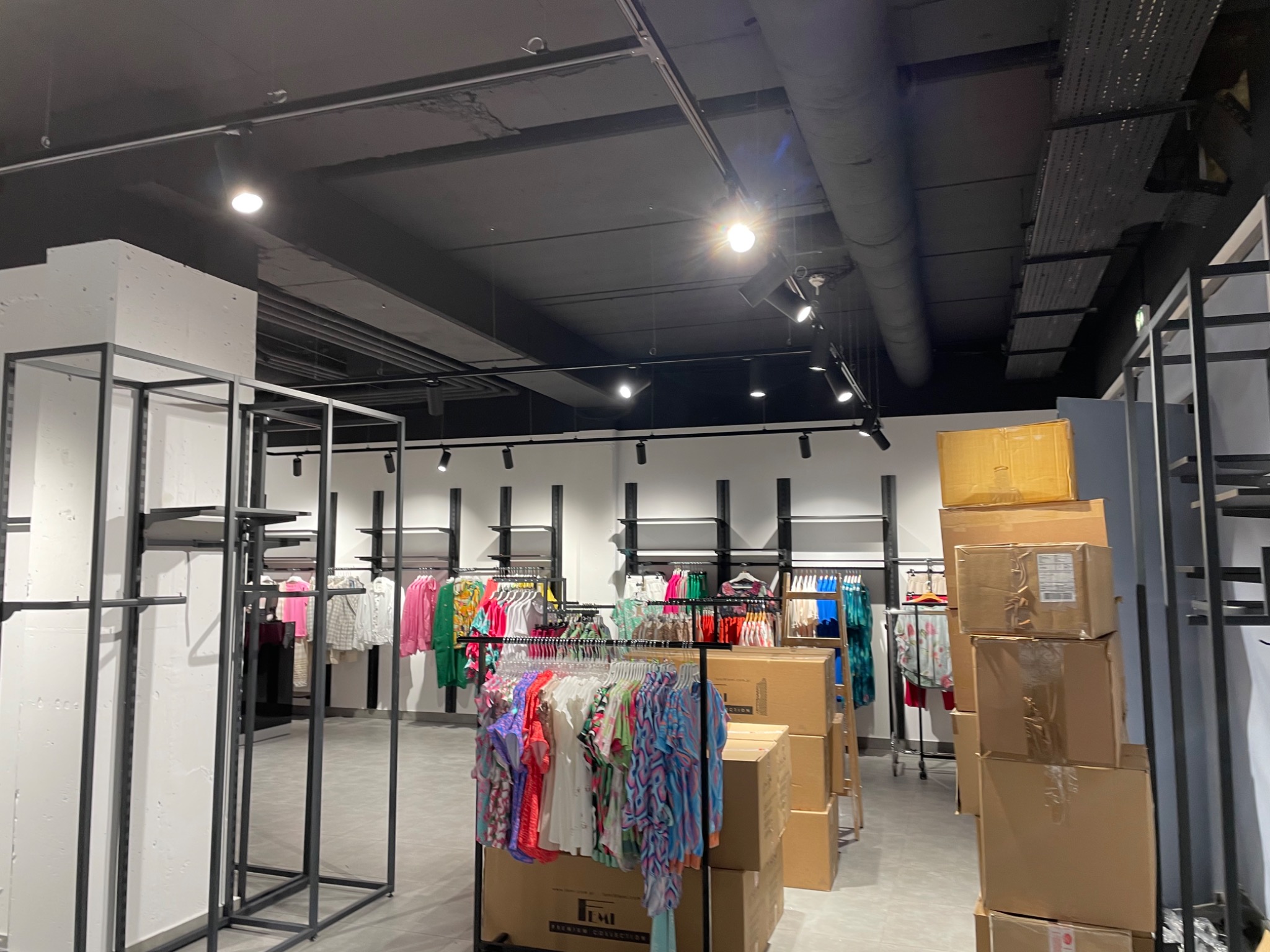 Wnętrze sklepu odzieżowego w trakcie aranżacji: widoczne regały, wieszaki z ubraniami oraz stos kartonowych pudeł, surowy beton na suficie z widocznymi instalacjami.