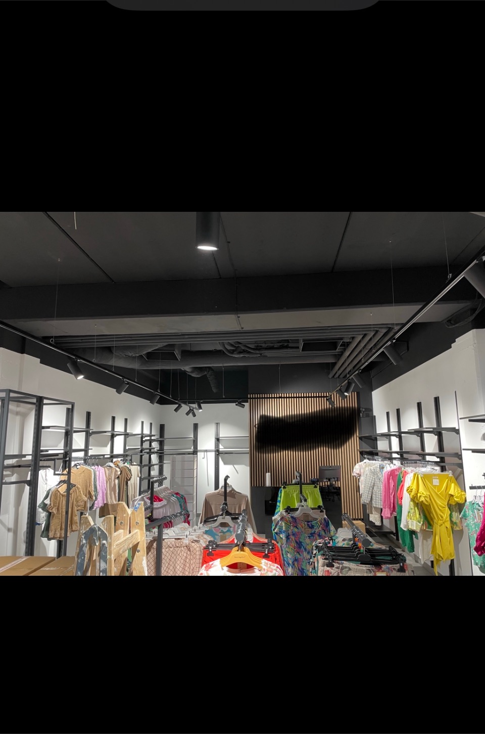 Wnętrze sklepu odzieżowego z czarnym sufitem, widoczne metalowe regały i wieszaki z ubraniami w różnych kolorach. Na ścianie dekoracyjna drewniana okładzina.