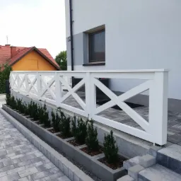 Biała, drewniana balustrada z krzyżowym wzorem przy schodach wejściowych do domu, z rzędem małych iglaków w betonowych donicach, na tle szarej kostki brukowej i elewacji budynku.