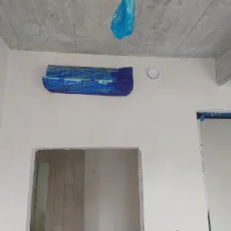 Klimatyzator zabezpieczony folią malarską na ścianie w surowym wnętrzu podczas remontu, widoczny otwór wentylacyjny i surowy betonowy strop.