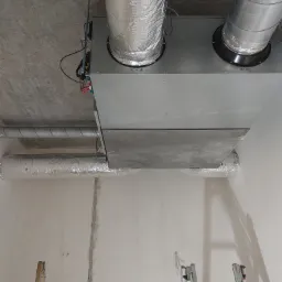 Widok z dołu na zamontowany system rekuperacji z widocznymi rurami wentylacyjnymi i przewodami elektrycznymi, w surowym stanie wykończenia wnętrza, z częściowo widoczną drabiną.