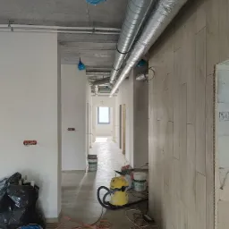 Wnętrze w trakcie wykańczania: widoczne rury wentylacyjne pod sufitem, ściany częściowo pomalowane i wykończone panelami, na podłodze narzędzia i worki na śmieci.