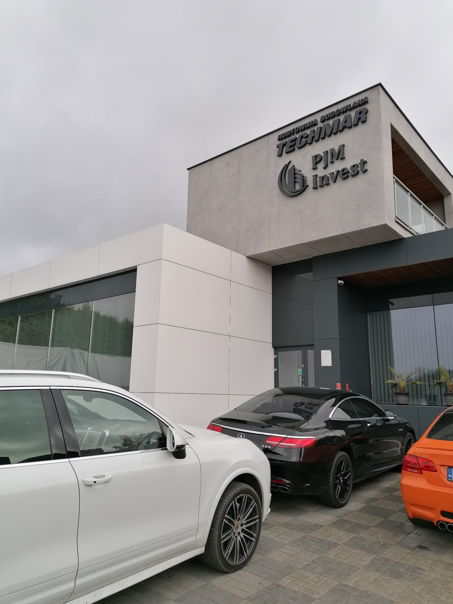 Nowoczesny budynek biurowy z logo firmy TECHMAR PJM Invest, z zaparkowanymi trzema samochodami: białym SUV-em, czarnym coupe Mercedes S63 i pomarańczowym BMW.