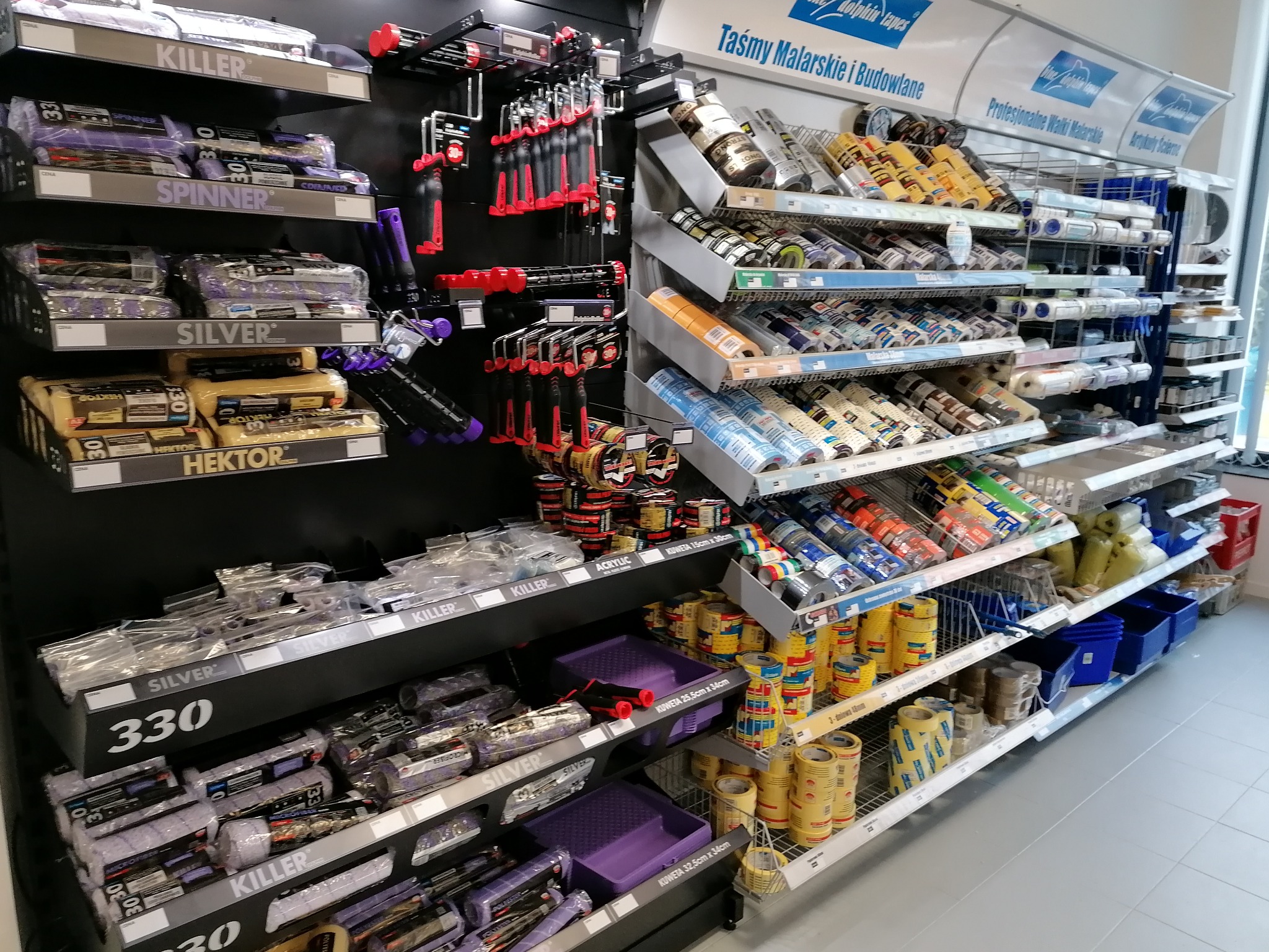 Ekspozycja w sklepie budowlanym: wałki malarskie Killer, Silver i Hektor, kuwety malarskie w odcieniach fioletu, taśmy malarskie Dolphin tapes oraz różnorodne narzędzia malarskie na czarnym tle.