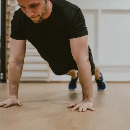 Mężczyzna w czarnej koszulce i sportowych butach wykonuje pompkę na drewnianej podłodze w jasnym pomieszczeniu z białymi panelami ściennymi.