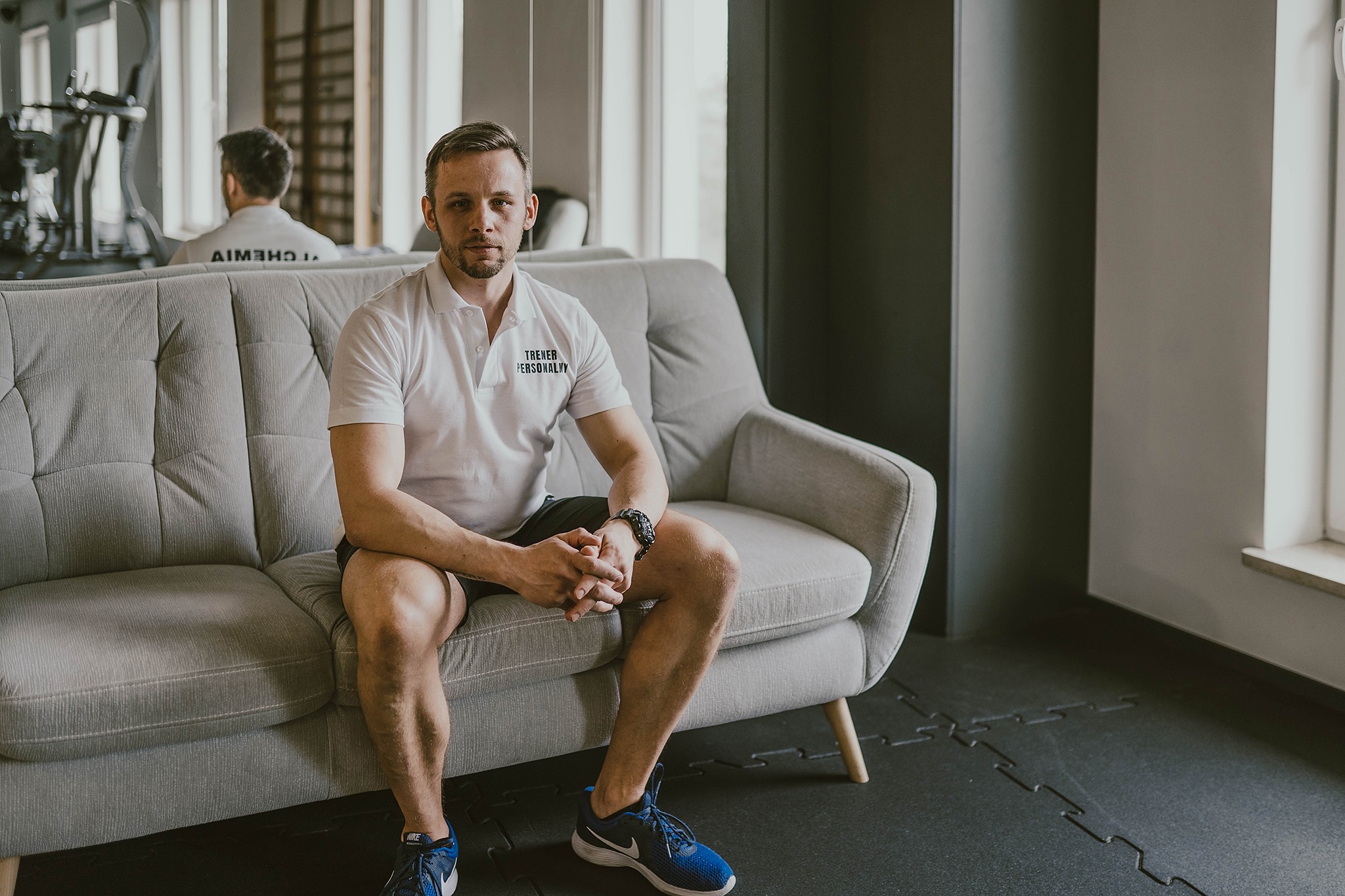 Mężczyzna w białej koszulce polo z napisem 'TRENER PERSONALNY' siedzi na szarej sofie w siłowni, za nim w lustrze odbicie ćwiczącego na maszynie.