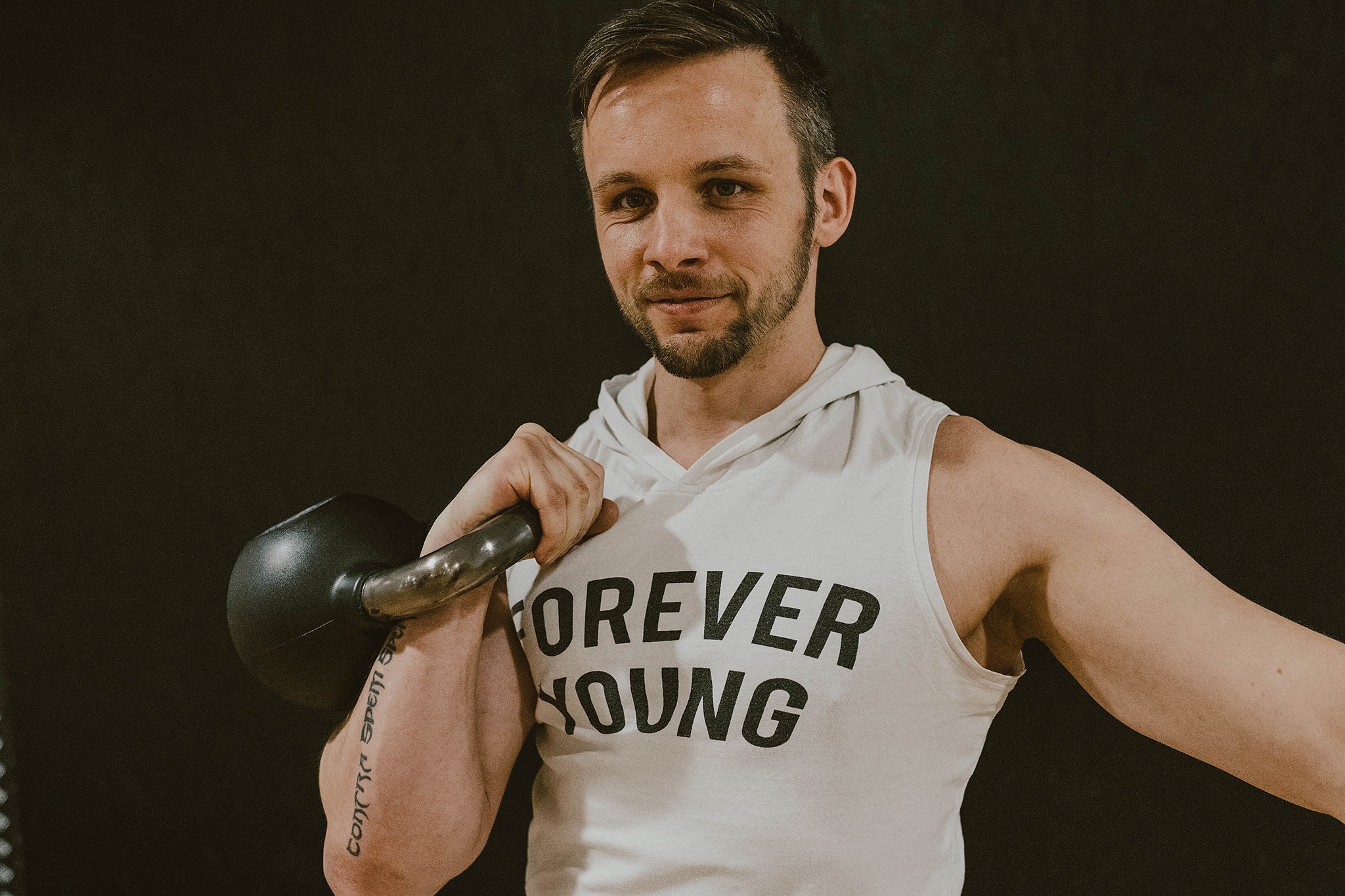 Uśmiechnięty mężczyzna w białej koszulce z napisem 'Forever Young' trzyma kettlebell, widoczny tatuaż na ramieniu.