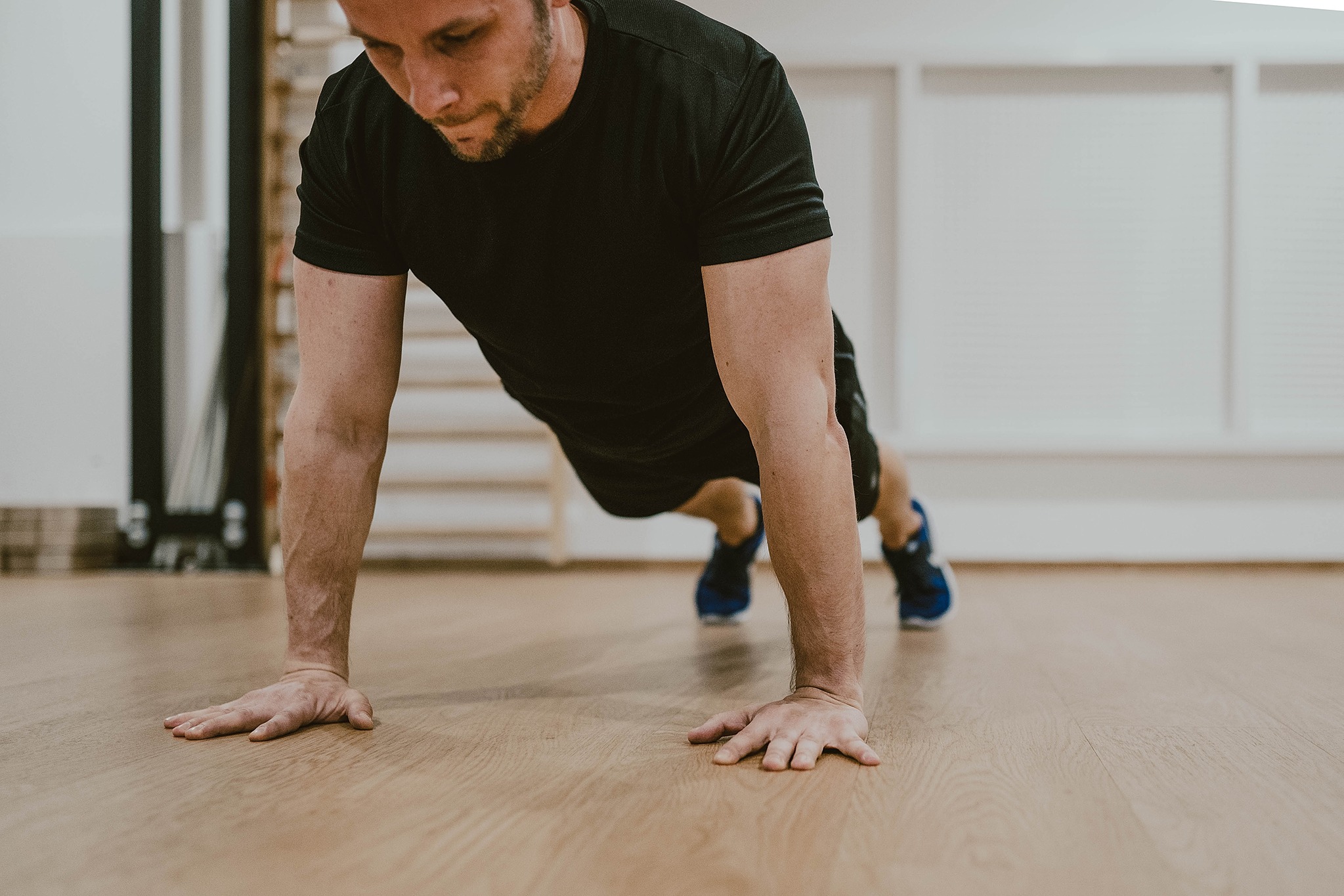 Mężczyzna w czarnej koszulce i sportowych butach wykonuje pompkę na drewnianej podłodze w jasnym pomieszczeniu z białymi panelami ściennymi.