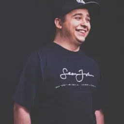 Uśmiechnięty mężczyzna w czarnej koszulce z napisem 'Sean John' i czapce z daszkiem na ciemnym tle.