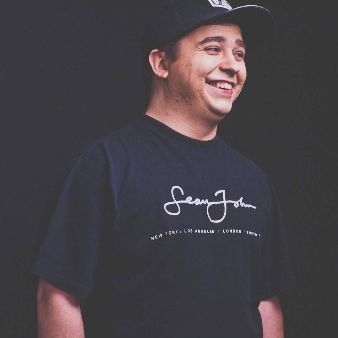 Uśmiechnięty mężczyzna w czarnej koszulce z napisem 'Sean John' i czapce z daszkiem na ciemnym tle.