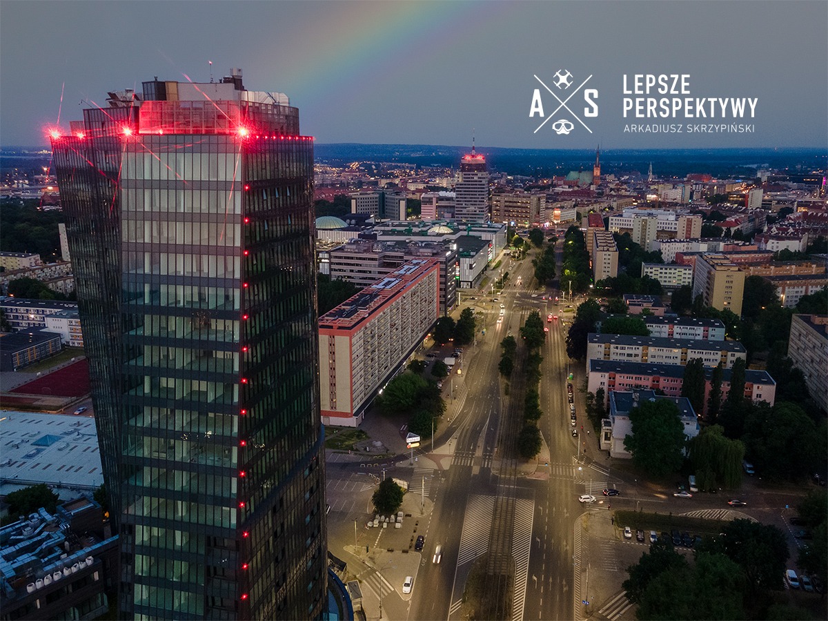 Nocny widok na miasto z drapaczem chmur oświetlonym czerwonymi punktami, tęcza na niebie, logo 'Lepsze Perspektywy Arkadiusz Skrzypiński' w prawym górnym rogu.