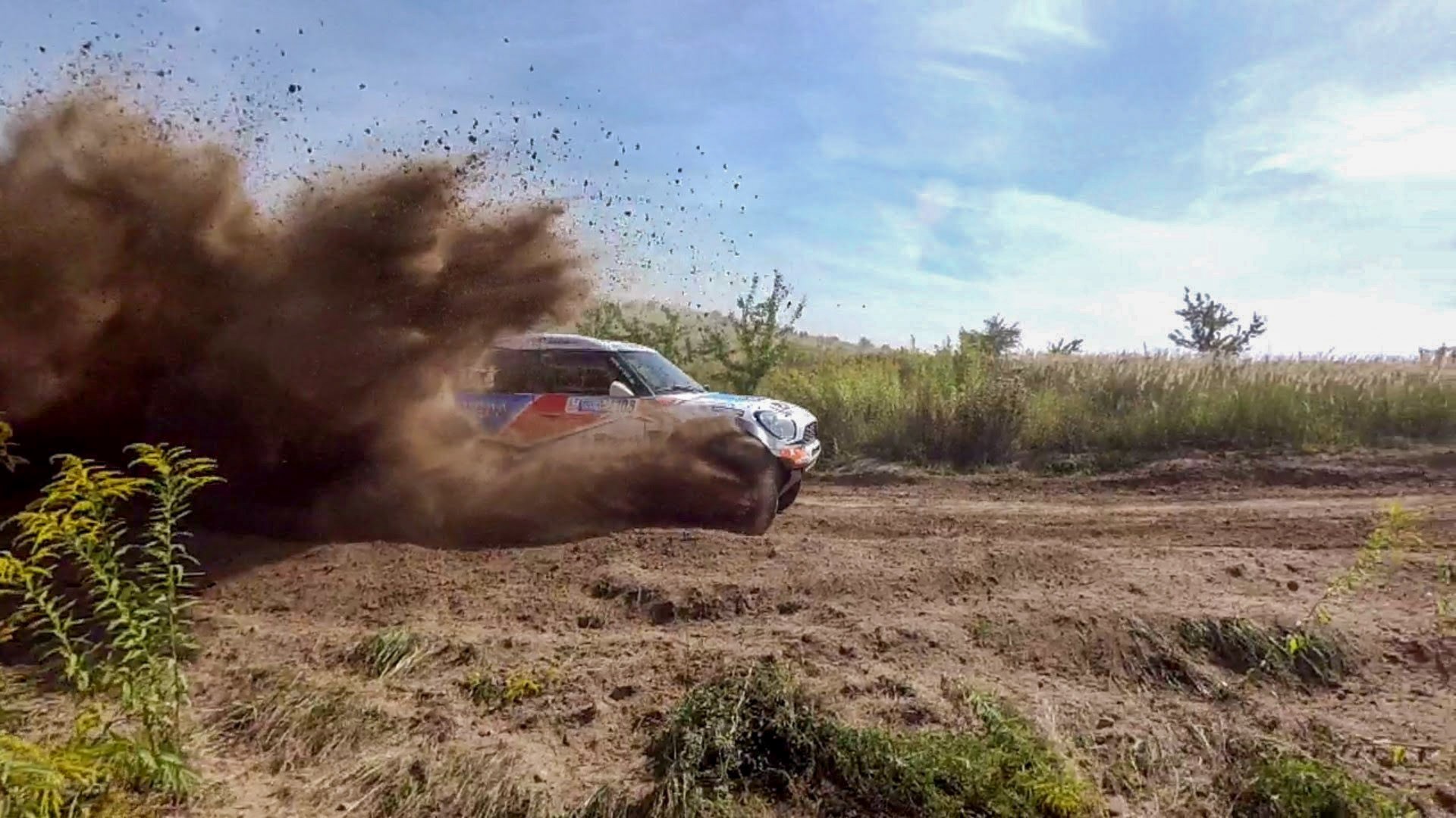 Rajdowy samochód Mini Cooper w trakcie dynamicznego przejazdu po piaszczystej trasie, spod kół wzbija się chmura kurzu i piasku.