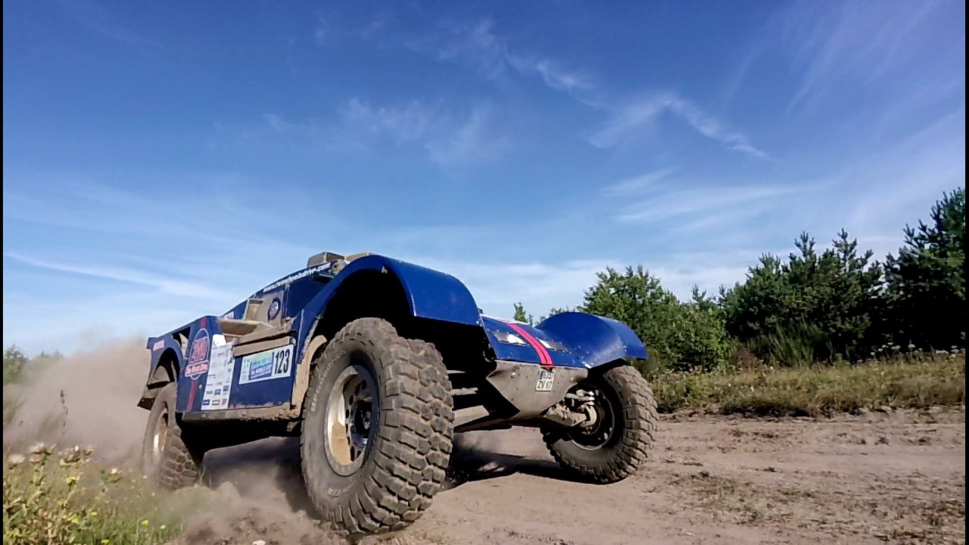 Niebieski samochód rajdowy z numerem 123 na boku, pędzący po piaszczystej drodze, z kurzem unoszącym się spod kół na tle błękitnego nieba i zieleni drzew.