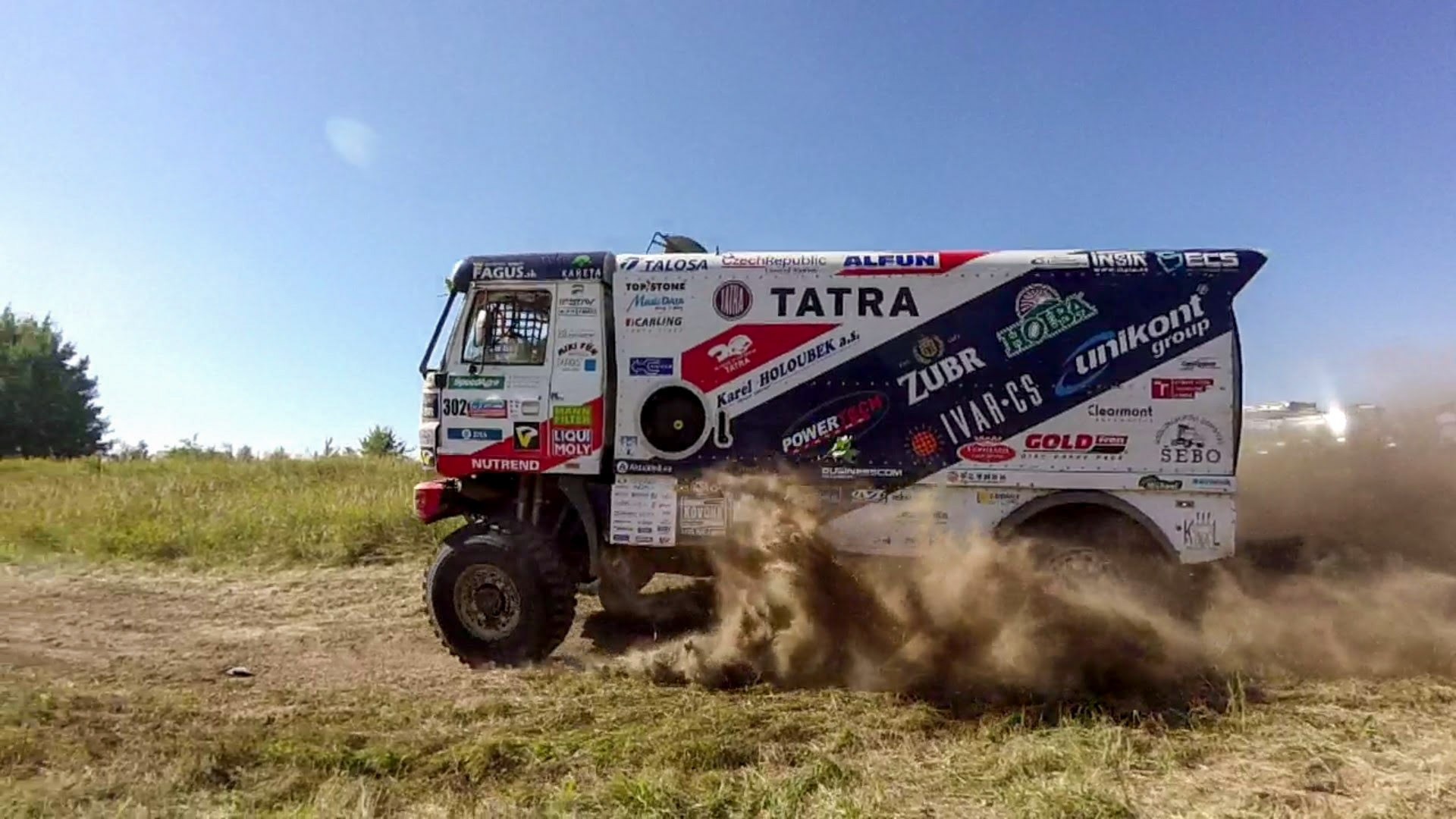 Ciężarówka rajdowa Tatra z licznymi logotypami sponsorów, pędząca po polnej drodze, wzbijająca tumany kurzu.