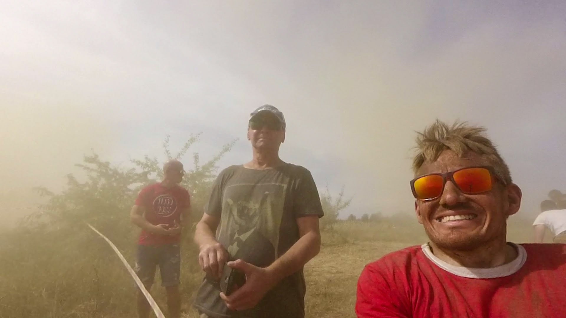 Mężczyzna w czerwonej koszulce i okularach przeciwsłonecznych, uśmiechnięty i pokryty błotem, na tle zakurzonego krajobrazu z innymi osobami w tle.