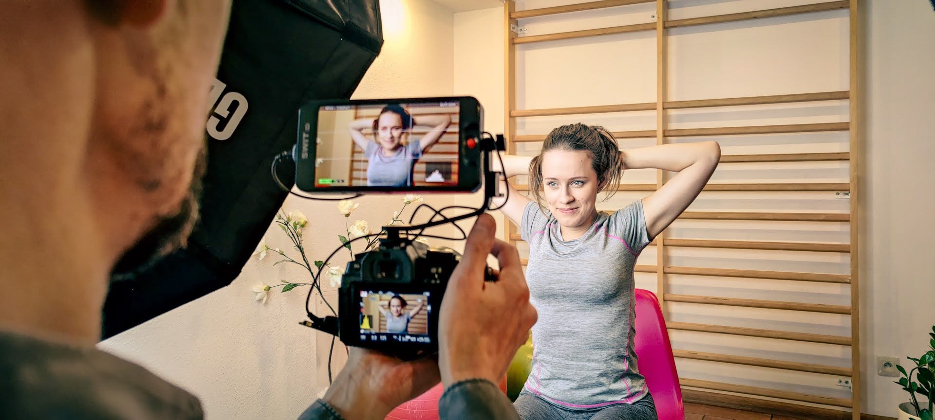 Kobieta pozuje podczas sesji zdjęciowej, widoczna przez obiektyw aparatu i ekran smartfona, z drabinką gimnastyczną w tle.