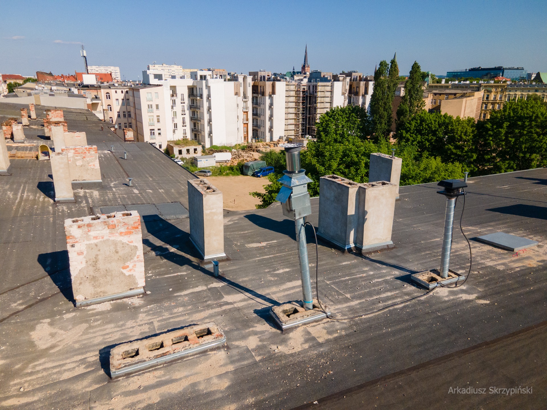 Widok z góry na płaski dach budynku z wystającymi kominami, wentylacją i instalacjami, w tle zabudowa miejska.