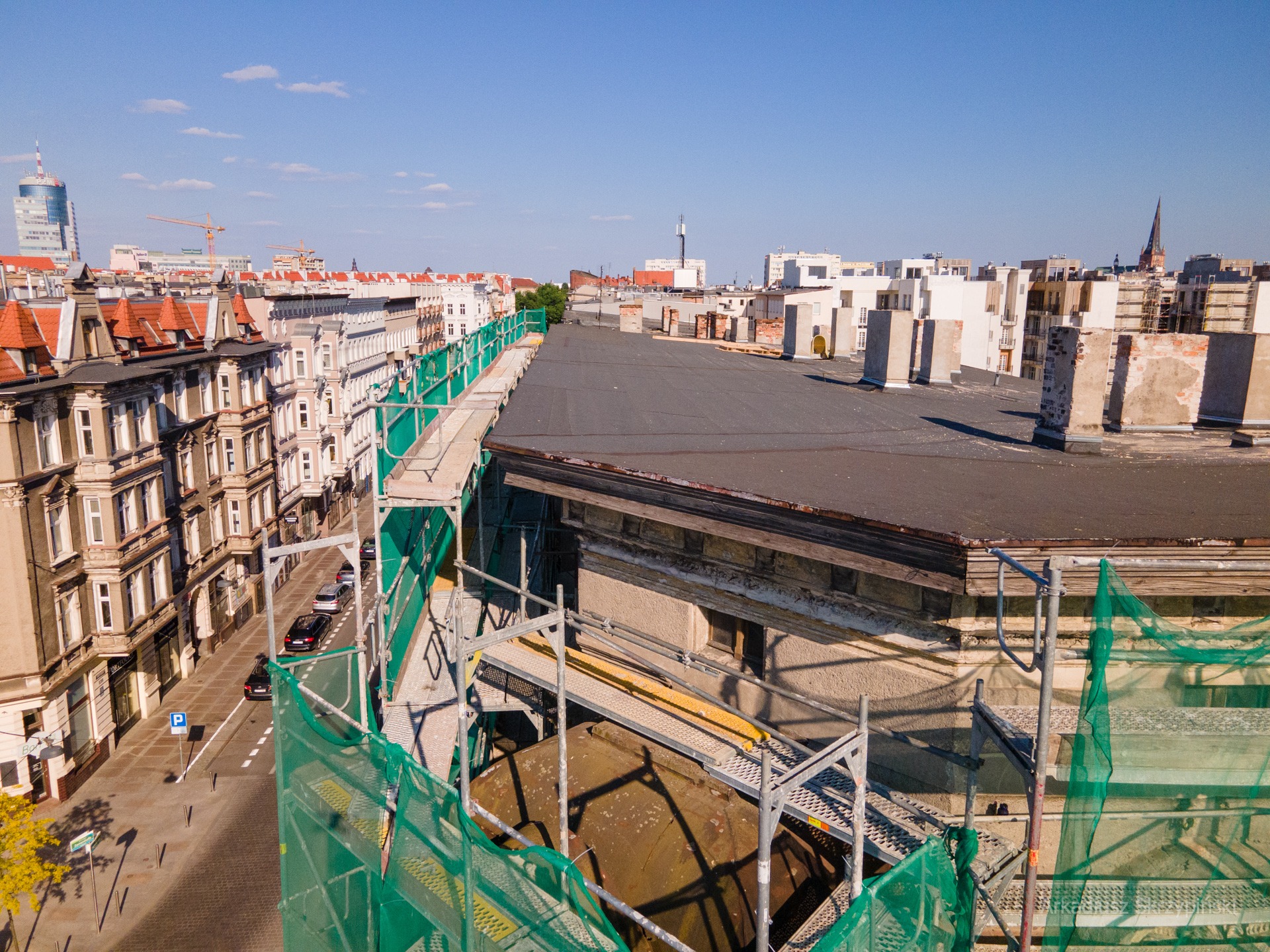 Widok z góry na kamienice w mieście, rusztowania z siatką zabezpieczającą na dachu budynku w trakcie renowacji, kominy w tle.