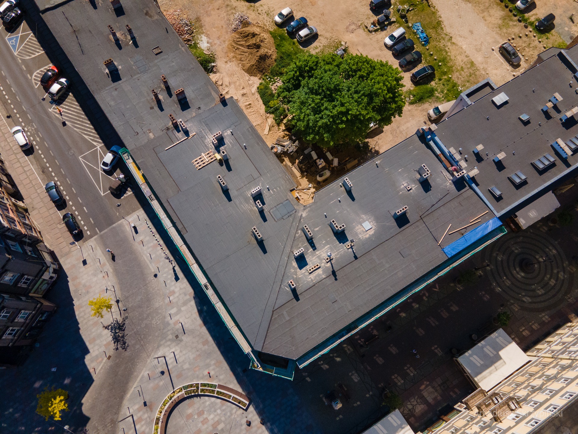 Widok z lotu ptaka na miejski krajobraz z budynkiem o płaskim dachu, parkingiem z autami i drzewem, widoczne elementy infrastruktury miejskiej.