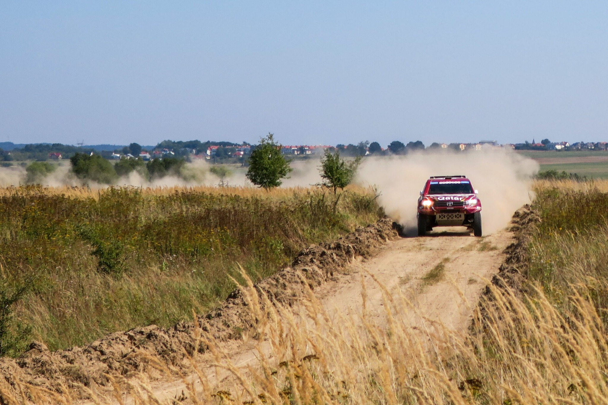 Czerwony samochód rajdowy z napisem Qatar pędzi po piaszczystej drodze, wzbijając tumany kurzu na tle wiejskiego krajobrazu z widocznymi zabudowaniami.