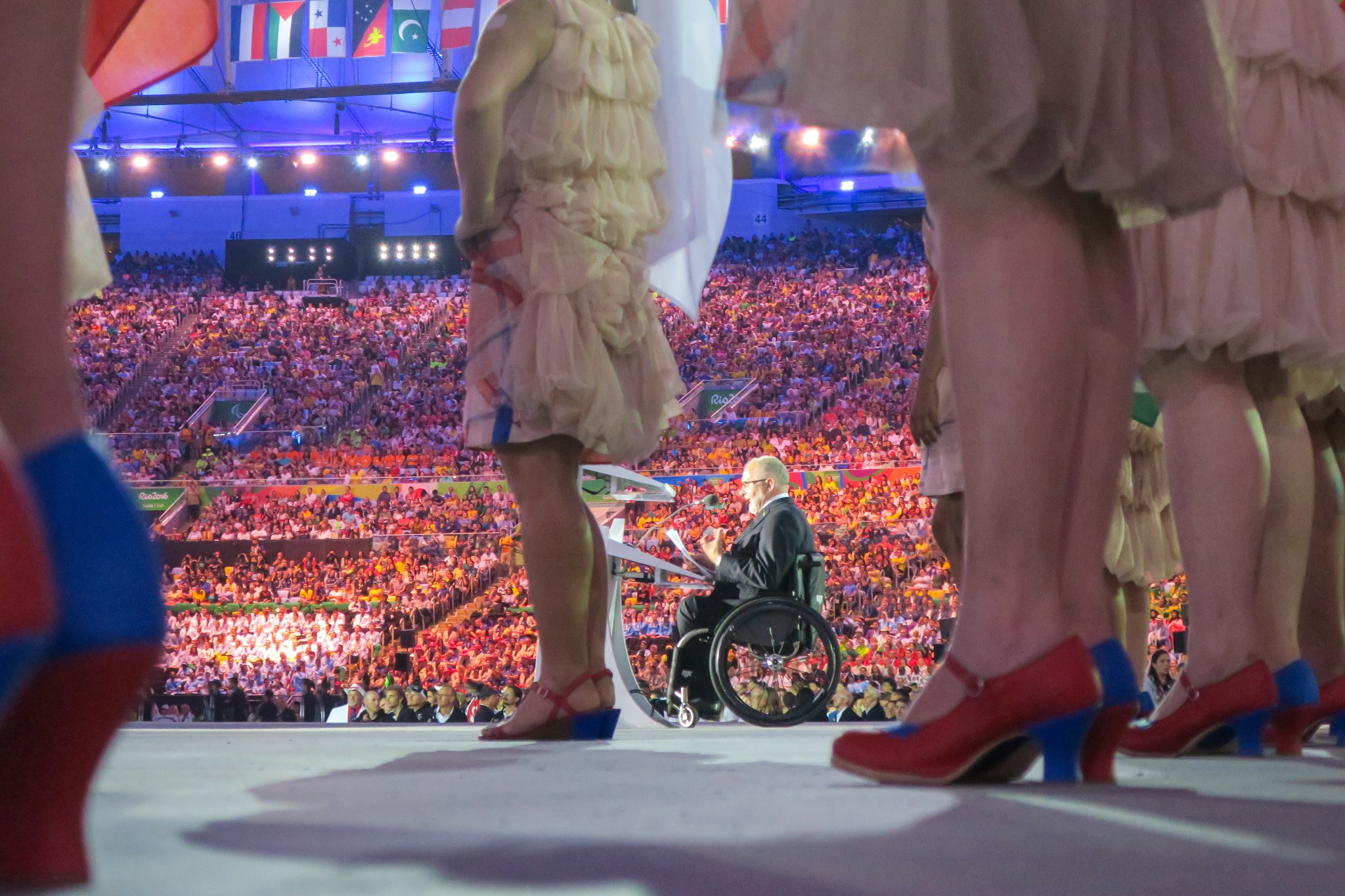 Uroczysta ceremonia otwarcia wydarzenia sportowego z tłumem widzów, tancerkami w strojach z falbankami i mówcą na wózku inwalidzkim.